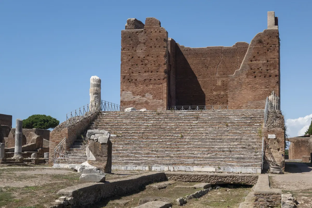 The Capitolium of Ostia Antica today from the forum: the flight of steps, the brick core of the temple without columns, on the left the column of the basilica, blue sky (photo)