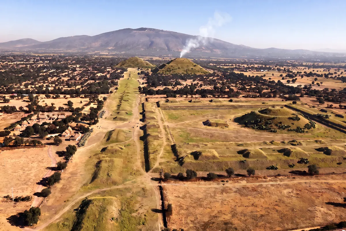 KI-Vision: Teotihuacán um 1500 aus der Luft, Sonnen- und Mondpyramide als grasbewachsene Hügel mit Nopalkakteen, die Straße der Toten als Reihe von Hügeln, Felder, ein kleines Dorf, Rauch von Räucherwerk auf der Sonnenpyramide